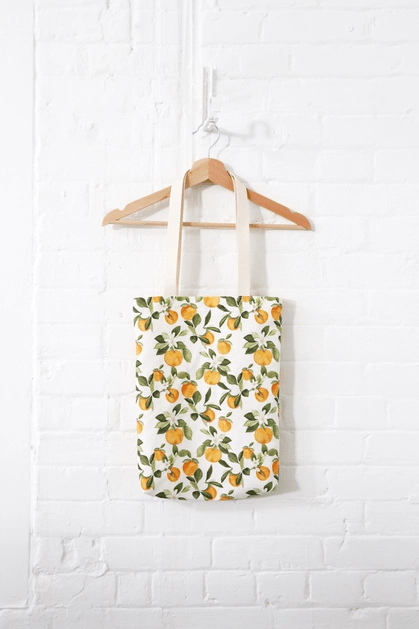 Tote bag in Clementine print, with various small clementine fruits, white flowers and green leaves, hung in wood hanger