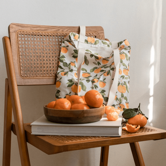 Tote bag in Clementine print, with various small clementine fruits, white flowers and green leaves, sitting on wood chair with book and bowl of clementines