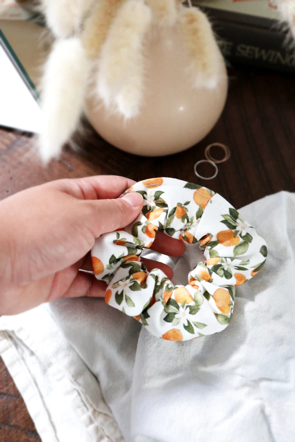 Female hand holding scrunchie in Clementine print, with various small clementine fruits, white flowers and green leaves